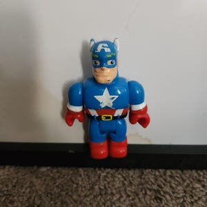 May include: A blue Captain America action figure with a white star on the chest, red gloves and boots, and a blue helmet with a white "A". The toy has a black belt and a tan face.