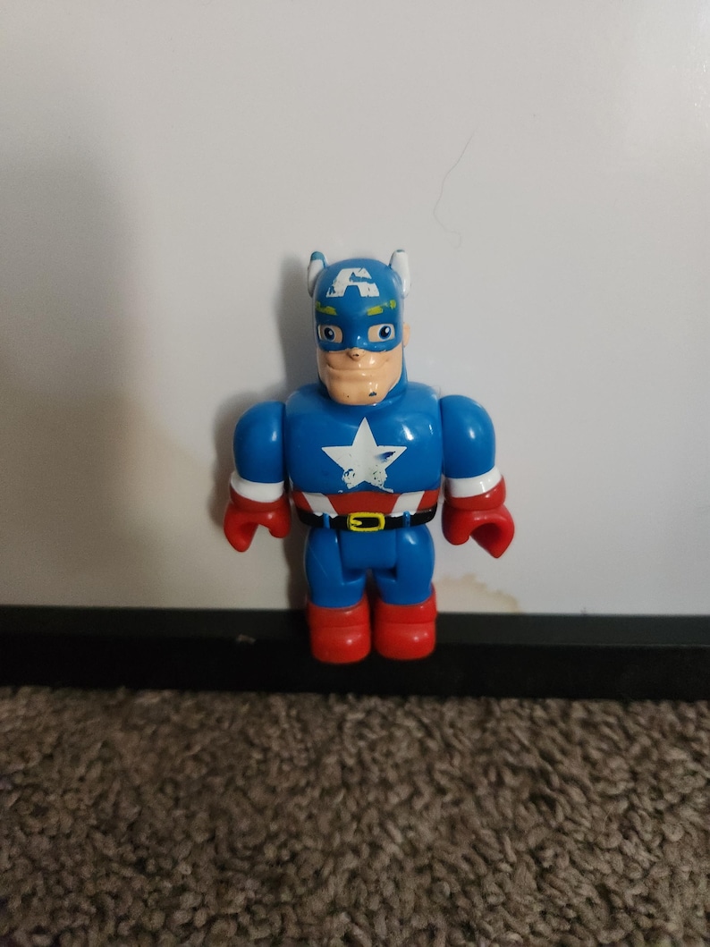 May include: A blue Captain America action figure with a white star on the chest, red gloves and boots, and a blue helmet with a white "A". The toy has a black belt and a tan face.