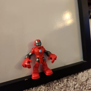 May include: A red and silver Iron Man action figure. The toy has a red helmet with a silver faceplate, red body, and silver arms. A blue circle is on the chest. The figure is standing on a carpeted surface.