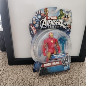 May include: A red and gold Iron Man action figure from the Marvel Avengers Assemble line. The figure is holding a blue repulsor blast effect piece. The packaging features a cartoon image of Iron Man, Captain America, Hulk, and Thor. The text on the packaging reads "Marvel Avengers Assemble" and "Repulsor Blast!"
