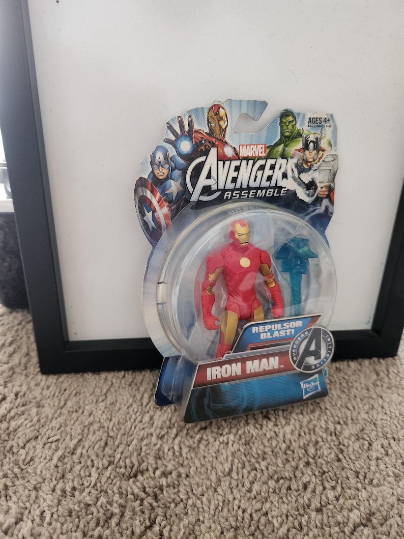 May include: A red and gold Iron Man action figure from the Marvel Avengers Assemble line. The figure is holding a blue repulsor blast effect piece. The packaging features a cartoon image of Iron Man, Captain America, Hulk, and Thor. The text on the packaging reads "Marvel Avengers Assemble" and "Repulsor Blast!"