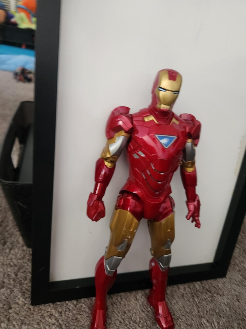 May include: An Iron Man action figure featuring a metallic red and gold suit with silver accents. The figure has a triangular blue arc reactor on the chest, a gold faceplate with white eye slits, and articulated joints throughout the body.