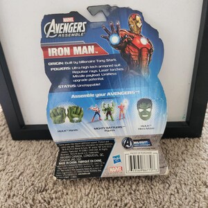 May include: A red, white, and blue card back for a Marvel Avengers Assemble Iron Man action figure. The card features a photo of Iron Man in his red and gold suit. The card also includes information about Iron Man's origin, powers, and status. The card also features a photo of Hulk's hands, a Mighty Battlers figure, and Hulk's hero mask.