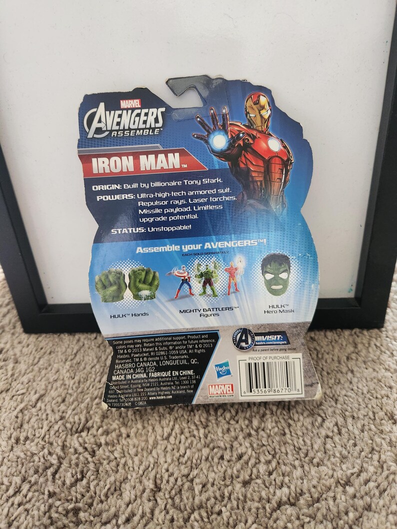 May include: A red, white, and blue card back for a Marvel Avengers Assemble Iron Man action figure. The card features a photo of Iron Man in his red and gold suit. The card also includes information about Iron Man's origin, powers, and status. The card also features a photo of Hulk's hands, a Mighty Battlers figure, and Hulk's hero mask.