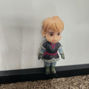 May include: A small figurine of a character with light orange hair, freckles, and brown eyes. The figure wears a dark gray tunic with a red belt, blue sleeves, and green gloves and boots. The figure is standing on a brown surface.