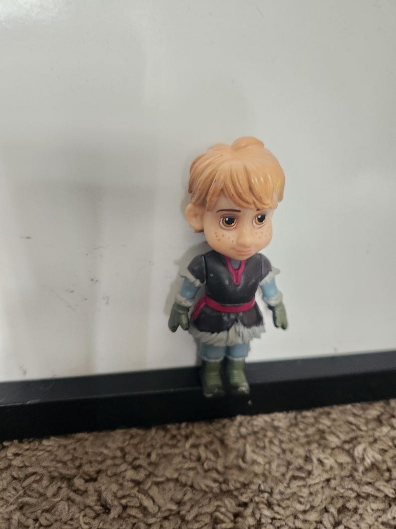 May include: A small figurine of a character with light orange hair, freckles, and brown eyes. The figure wears a dark gray tunic with a red belt, blue sleeves, and green gloves and boots. The figure is standing on a brown surface.