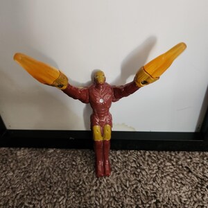 May include: A plastic Iron Man action figure in a dynamic pose, with arms outstretched and yellow energy blasts. The figure is primarily red and gold, with detailed armor features. The background is a white wall and brown carpet.