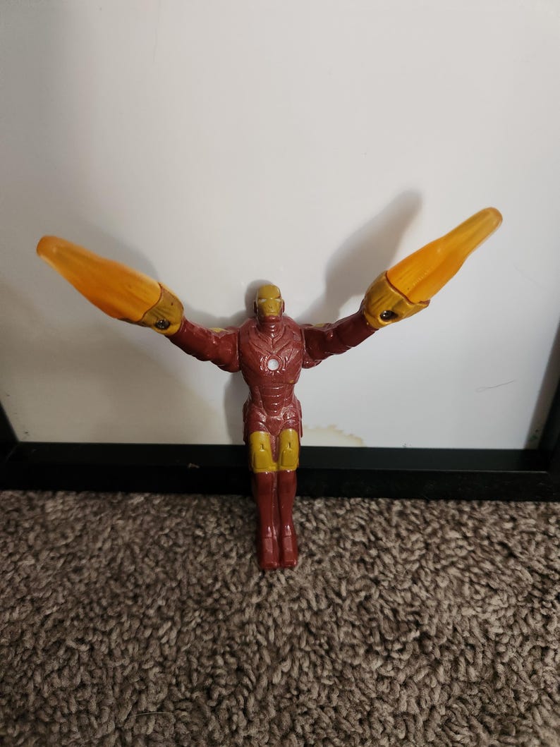 May include: A plastic Iron Man action figure in a dynamic pose, with arms outstretched and yellow energy blasts. The figure is primarily red and gold, with detailed armor features. The background is a white wall and brown carpet.
