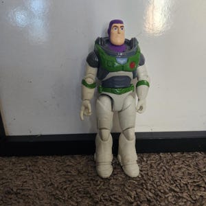 May include: A Buzz Lightyear action figure, featuring a white and green spacesuit with purple accents. The toy has a light skin tone and a determined expression. The figure is standing on a brown carpet.
