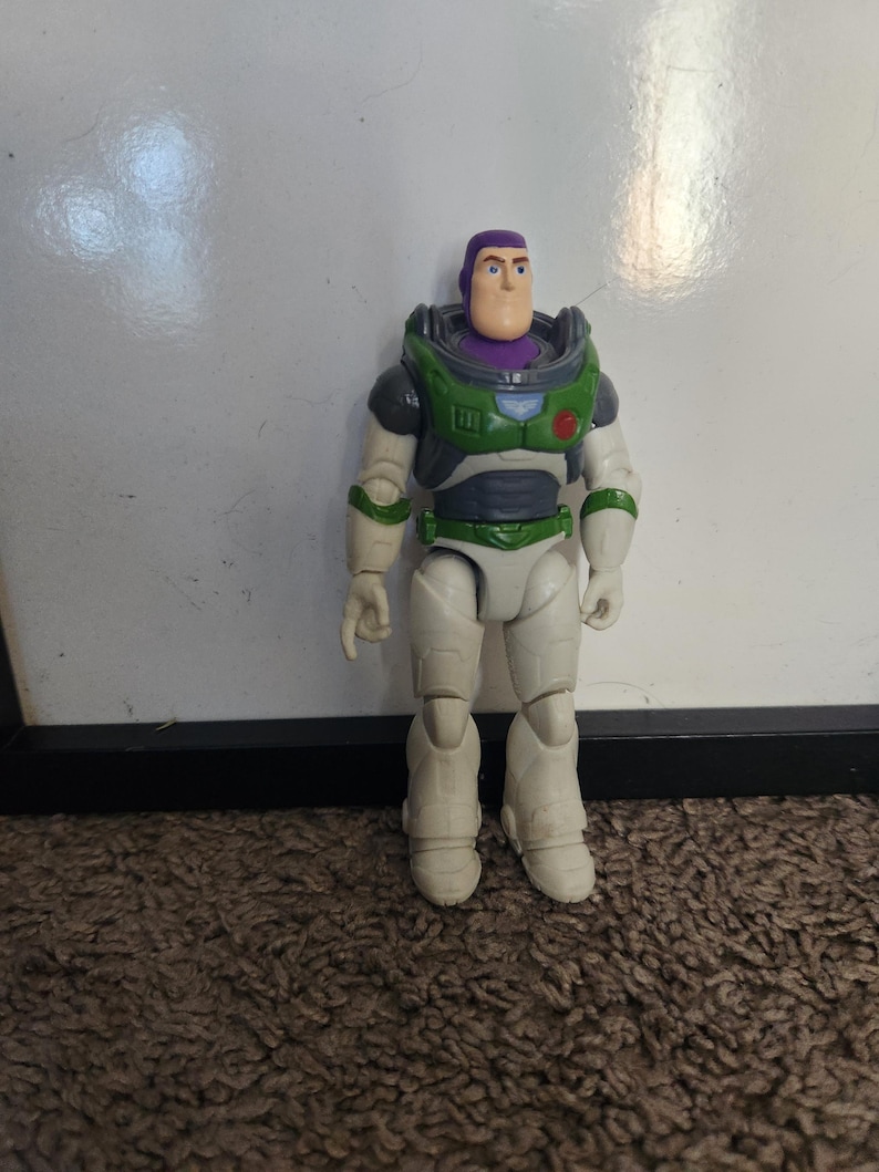 May include: A Buzz Lightyear action figure, featuring a white and green spacesuit with purple accents. The toy has a light skin tone and a determined expression. The figure is standing on a brown carpet.