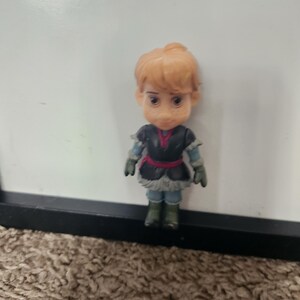 May include: A small figurine of a character with light brown hair styled in a bun. The figure wears a black and blue outfit with a red belt and green boots. The figurine is standing in front of a white background.