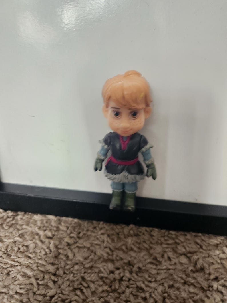 May include: A small figurine of a character with light brown hair styled in a bun. The figure wears a black and blue outfit with a red belt and green boots. The figurine is standing in front of a white background.