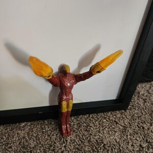 May include: A red and gold Iron Man action figure with outstretched arms and yellow energy blasts. The figure is standing in front of a white surface and a black frame. The toy is a collectible item.