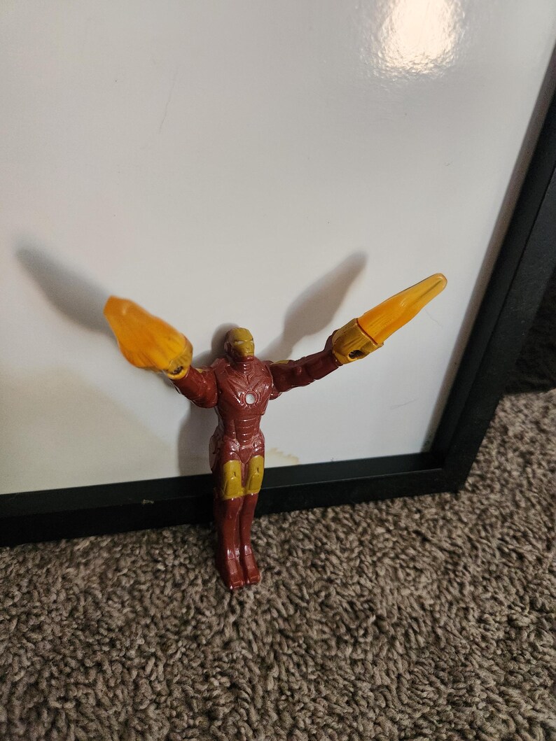 May include: A red and gold Iron Man action figure with outstretched arms and yellow energy blasts. The figure is standing in front of a white surface and a black frame. The toy is a collectible item.