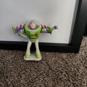 Puede incluir: Una figura de acci&oacute;n de Buzz Lightyear, con un traje espacial blanco y verde con detalles de alas rojas y blancas. La figura tiene un casco transparente y est&aacute; posando con un brazo extendido. El juguete est&aacute; sobre una peque&ntilde;a base blanca.