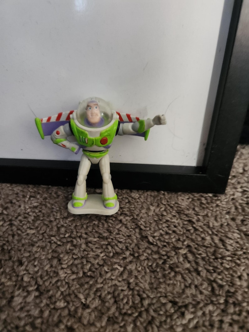 Puede incluir: Una figura de acci&oacute;n de Buzz Lightyear, con un traje espacial blanco y verde con detalles de alas rojas y blancas. La figura tiene un casco transparente y est&aacute; posando con un brazo extendido. El juguete est&aacute; sobre una peque&ntilde;a base blanca.