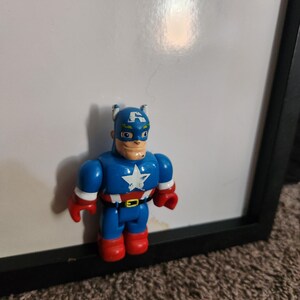 May include: A Captain America action figure. The figure is primarily blue with red gloves and boots. The chest features a white star on a white background. The figure has a blue mask with a white "A" on the forehead.