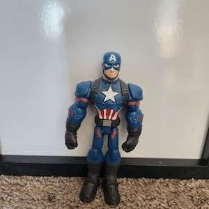 May include: A Captain America action figure. The figure is primarily blue with red and white accents. It features a white star on the chest and a blue helmet with a white "A". The figure is standing on a brown carpet.
