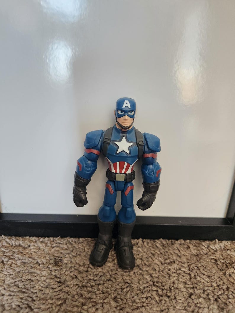 May include: A Captain America action figure. The figure is primarily blue with red and white accents. It features a white star on the chest and a blue helmet with a white "A". The figure is standing on a brown carpet.