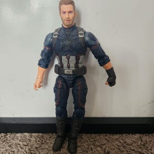 May include: A Captain America action figure. The figure is wearing a blue and black suit with a silver and white chest plate and a black belt. The figure has a detailed face and is standing on a brown carpet.