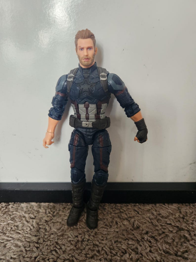 May include: A Captain America action figure. The figure is wearing a blue and black suit with a silver and white chest plate and a black belt. The figure has a detailed face and is standing on a brown carpet.