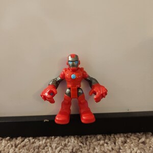 May include: A red and silver Iron Man action figure. The toy has a red body, legs, and hands, with silver arms and a helmet. The figure has a blue circle on its chest and blue eyes.