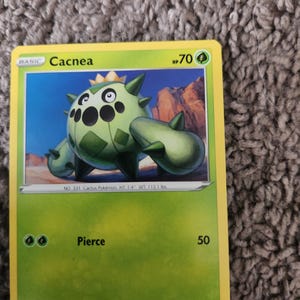 May include: A Cacnea Pokémon trading card with a green and yellow border. The card features a green cactus-like creature with black eyes and spikes. The card text includes the name "Cacnea", HP 70, and the attack "Pierce" for 50.