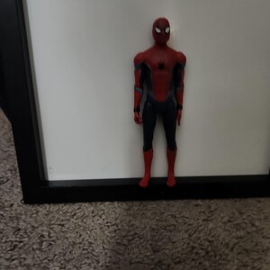 Op de afbeelding: Een Spider-Man actiefiguur, met een rood en zwart pak en een gedetailleerd masker. De figuur staat op een tapijt, met een witte achtergrond en een zwart frame. De figuur is ongeveer 15 cm hoog.