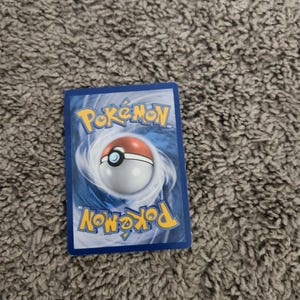 May include: A blue Pokémon trading card with the Pokémon logo and a Poké Ball design on the back. The card is rectangular and has a glossy finish. The background is a textured, gray surface.