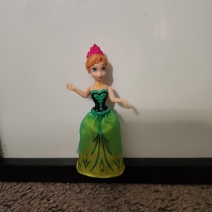 May include: A small princess doll with red hair and a pink crown. She is wearing a green and yellow dress with a black bodice. The doll is positioned against a white backdrop.