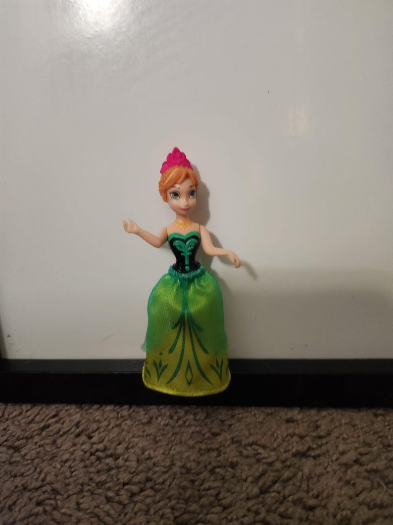 May include: A small princess doll with red hair and a pink crown. She is wearing a green and yellow dress with a black bodice. The doll is positioned against a white backdrop.