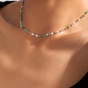 May include: A delicate beaded necklace featuring a sequence of small, round beads in alternating shades of light green, white, and metallic gold. The necklace rests against the neck, showcasing a simple, thin chain design with evenly spaced beads.