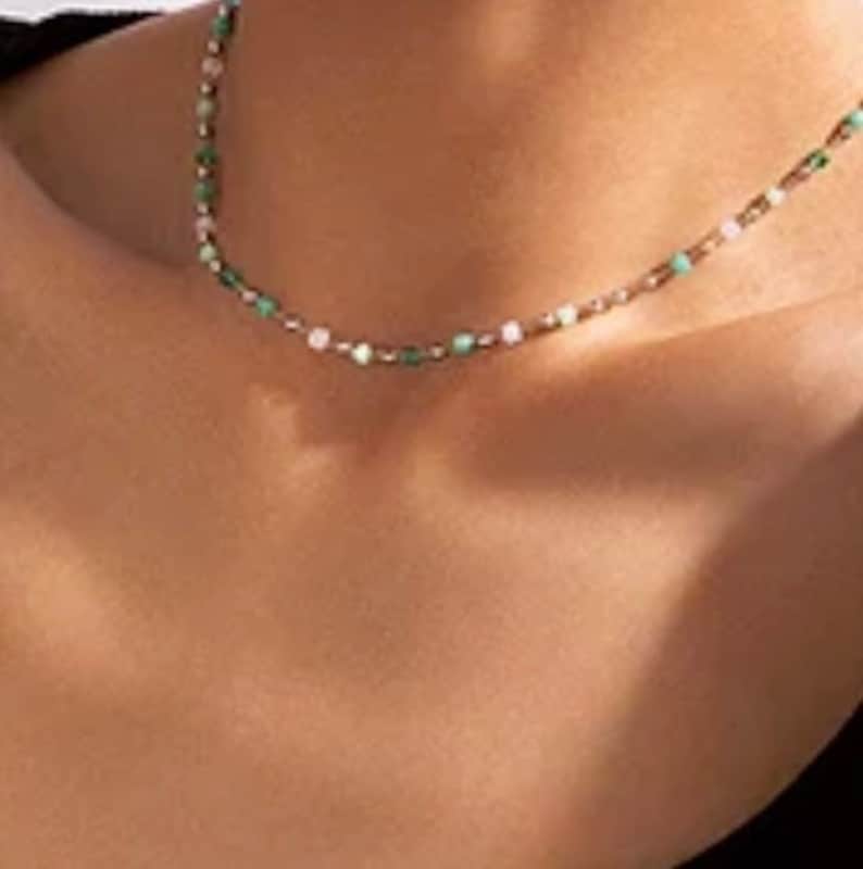 May include: A delicate beaded necklace featuring a sequence of small, round beads in alternating shades of light green, white, and metallic gold. The necklace rests against the neck, showcasing a simple, thin chain design with evenly spaced beads.
