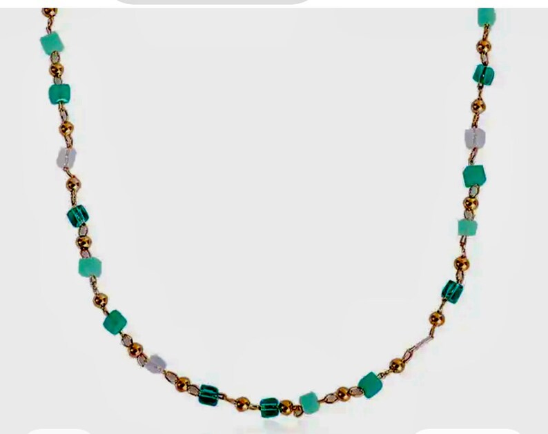 May include: A necklace featuring a delicate gold-toned chain interspersed with small, polished gold-colored spherical beads and various square-shaped beads in shades of light green, teal, and translucent white, arranged in a repeating pattern.