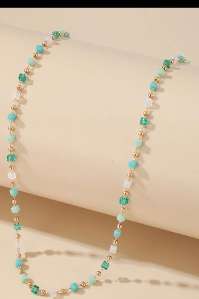May include: A delicate necklace featuring a thin gold-tone chain interspersed with small, square-shaped beads in shades of light green, dark green, and translucent white, alternating with tiny, round, polished gold-tone metal spacer beads.