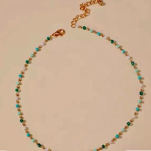 May include: A delicate beaded necklace featuring a repeating pattern of small gold-toned metallic spheres, translucent white beads, and square-shaped beads in shades of turquoise and emerald green, finished with a gold-toned lobster clasp and chain.
