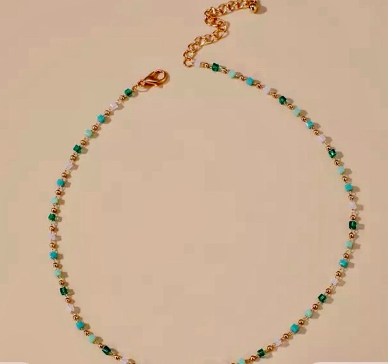 May include: A delicate beaded necklace featuring a repeating pattern of small gold-toned metallic spheres, translucent white beads, and square-shaped beads in shades of turquoise and emerald green, finished with a gold-toned lobster clasp and chain.