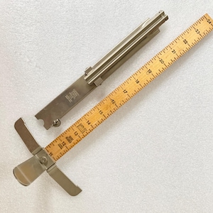 May include: A silver metal measuring tool with a ruler attached. The ruler is marked in inches from 10 to 24. The tool is used for measuring fabric.