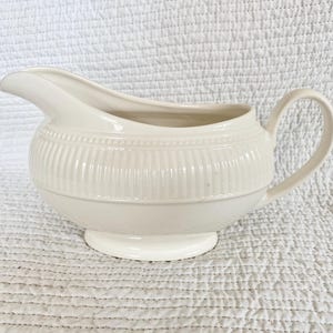 May include: Cream-colored gravy boat with a rounded body and a small, circular base. The boat features a spout, a handle, and vertical ribbed detailing around the middle. The gravy boat is made of ceramic.
