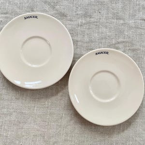 May include: Two off-white ceramic saucers with a central indentation and a raised rim. The word "SAUCER" is printed in navy blue on each saucer. The saucers are placed on a textured, light gray surface.