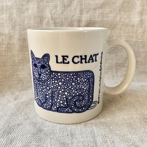 May include: A white ceramic mug with a blue cat illustration and the text "LE CHAT". The cat design is filled with small dots. The mug has a curved handle and is set against a neutral background.