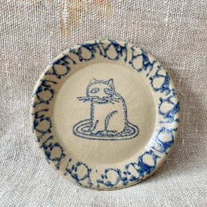 Puede incluir: Plato pequeño y redondo de cerámica, base crema con una ilustración de gato azul en el centro. El borde del plato presenta un patrón azul repetitivo. El gato está sentado sobre un óvalo azul.