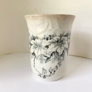May include: A white ceramic cup with a black and white floral design. The cup has a scalloped rim and a slightly flared base.