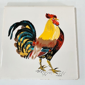 May include: A white ceramic tile with a colorful illustration of a rooster. The rooster is yellow, red, brown, black, and blue. The rooster is facing right and has its head turned to the left. The rooster is standing on a white background with black dots.