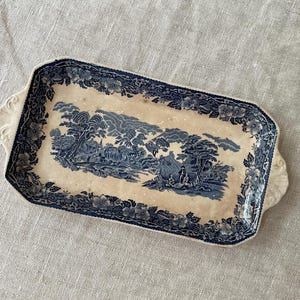 May include: A rectangular ceramic serving tray featuring a cream-colored base with a dark blue transferware pattern. The border displays a floral motif, while the center depicts a landscape with trees, hills, and a small figure.