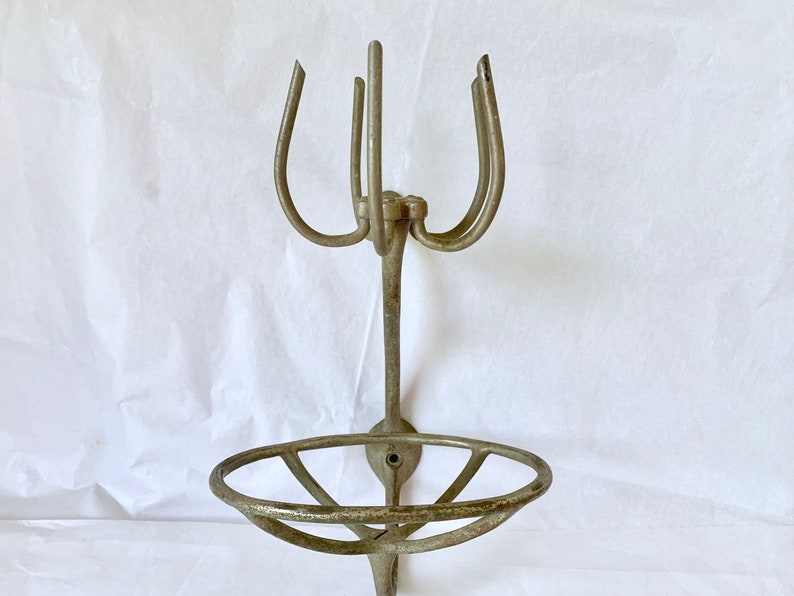 Puede incluir: Un jabonera y toallero de metal antiguo montado en la pared. La jabonera tiene un dise&ntilde;o circular y abierto, y el toallero tiene tres brazos curvos. El metal tiene un aspecto envejecido.
