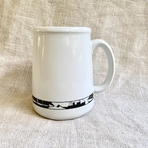 May include: White ceramic mug with a black graphic band featuring a mountain and boat design. The mug has a curved handle and a slightly flared rim. The mug is set against a neutral linen background.