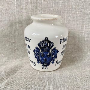 May include: White ceramic vase with blue and black decorative elements. The vase features a crown and fleur-de-lis design, along with text in a script font. The vase has a slightly distressed appearance, with a small base and a wide opening.
