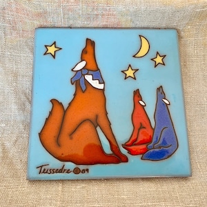 May include: Square ceramic tile with a light blue background featuring a howling wolf with a blue and white scarf, and two smaller wolves. The design includes a crescent moon and stars. The tile is signed "Tussedre © 09".