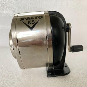 May include: A silver and black X-ACTO KS pencil sharpener. The sharpener has a silver metal body with the text "X-ACTO KS" and a black handle and base. The sharpener is designed to sharpen pencils.